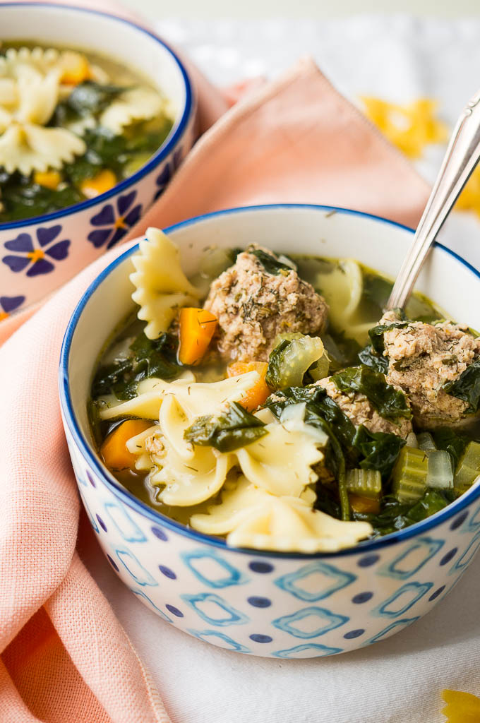 Italian Wedding Soup with turkey meatballs, fresh dill, and bowtie pasta  in a blue and white bowl on a peach napkin and a white tablecloth. Dried bowtie pasta.