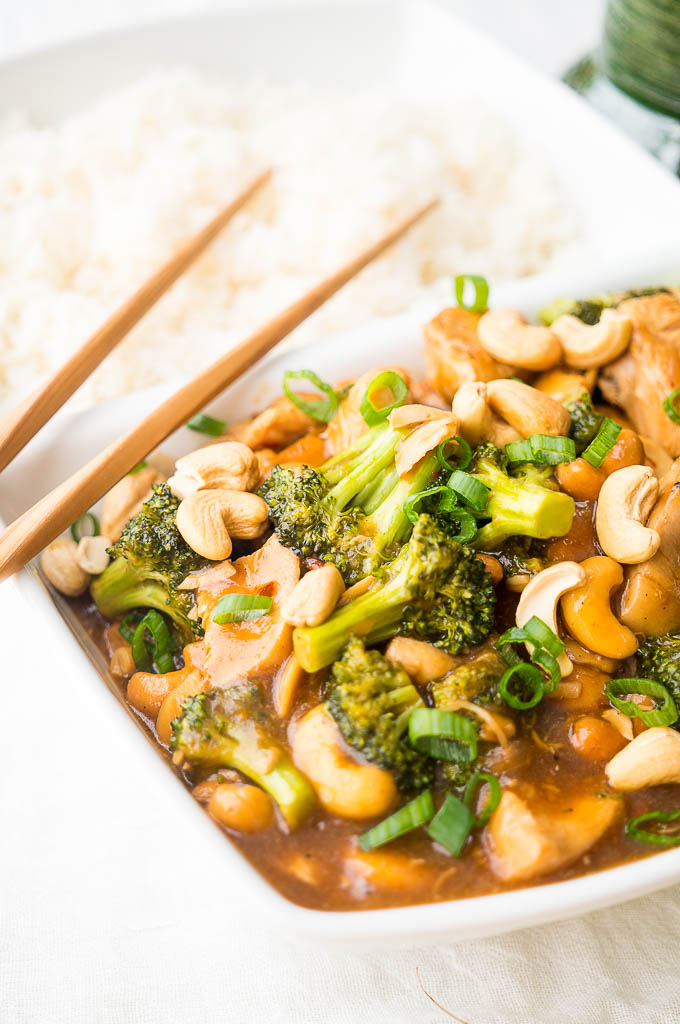 Cashew Chicken with Broccoli topped with green onions, and white rice in a white dish with wooden chopsticks on a white background. A green mug with copper utensils.