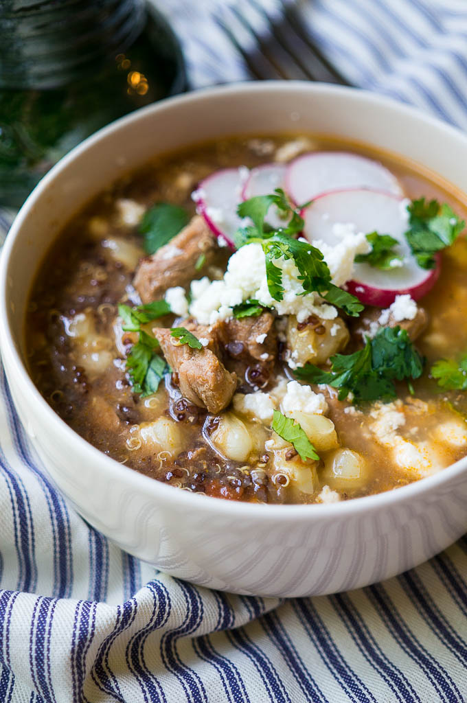 Simple Pork Posole in a white bowl, topped with fresh radishes, cilantro, and queso fresco on a blue and white striped napkin with a copper fork and a green mug.
