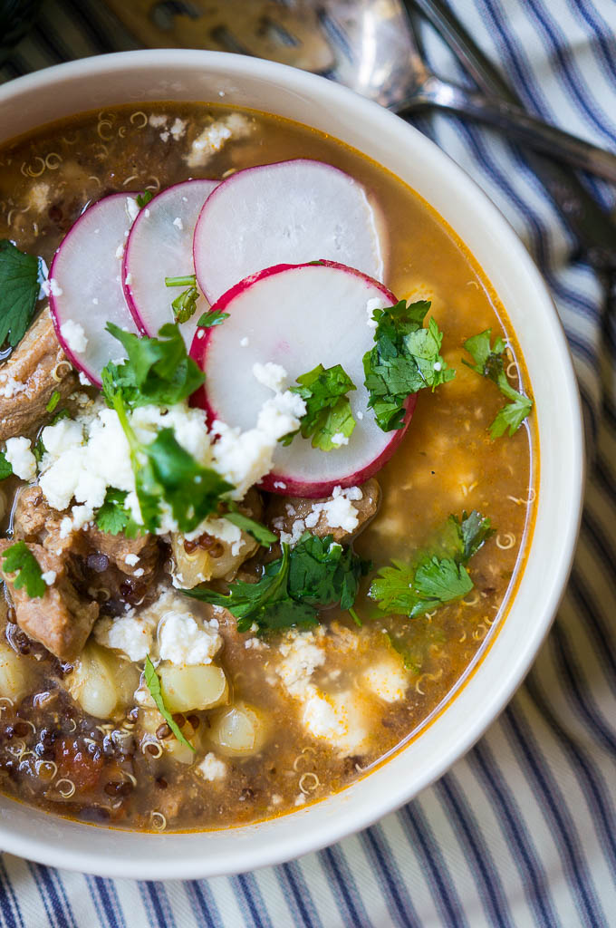 Simple pork posole in a white bowl, topped with fresh radishes, cilantro and queso fresco on a blue-and-white striped napkin with a copper spoon and fork and a green cup.