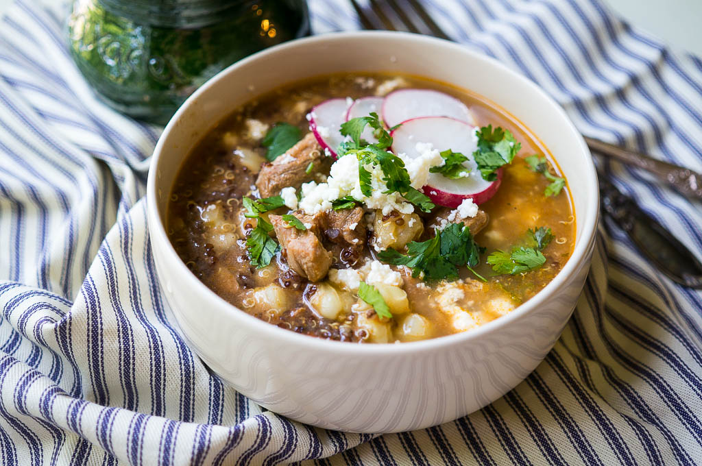 Simple Pork Posole in a white bowl, topped with fresh radishes, cilantro, and queso fresco on a blue and white striped napkin with a copper spoon and fork and a green mug.