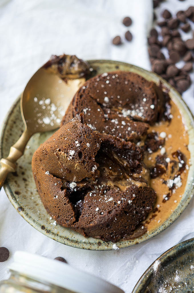 Dark Chocolate Almond Butter Lava Cake dusted with powdered sugar on a speckled ceramic plate with a copper spoon on a white tablecloth and chocolate chips.