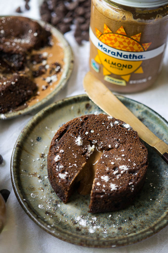 Dark Chocolate Almond Butter Lava Cakes dusted with powdered sugar on a speckled ceramic plate with a copper knife on a white tablecloth. Jar of Maranatha Almond butter and chocolate chips.