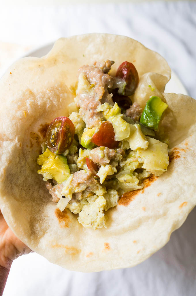 Sausage and egg breakfast taco topped with avocado and cherry tomatoes in a white tortilla on a white background.