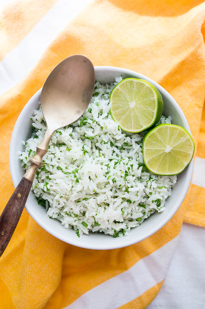 Cilantro Rice topped with lime wedges and a copper and wooden spoon in a white bowl on a yellow napkin.