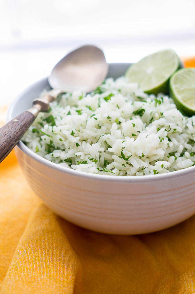 Cilantro Rice topped with lime wedges and a copper and wooden spoon in a white bowl on a yellow napkin.