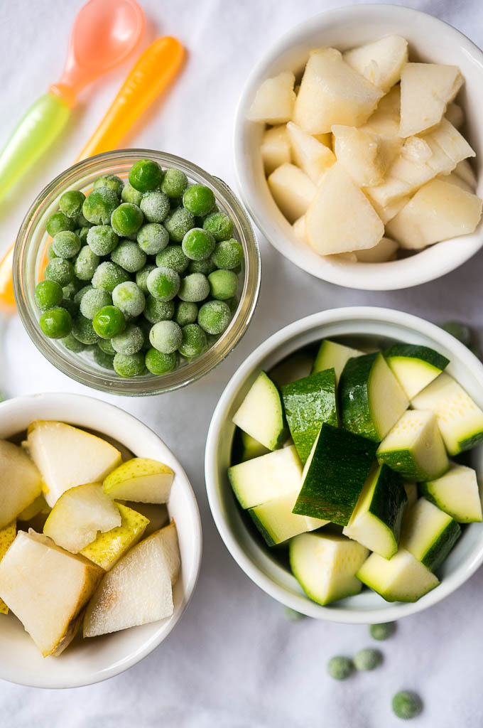 Frozen peas in a glass bowl, pears in a white bowl, zucchini in a white bowl, apples in a white bowl, and baby spoon on a white background.