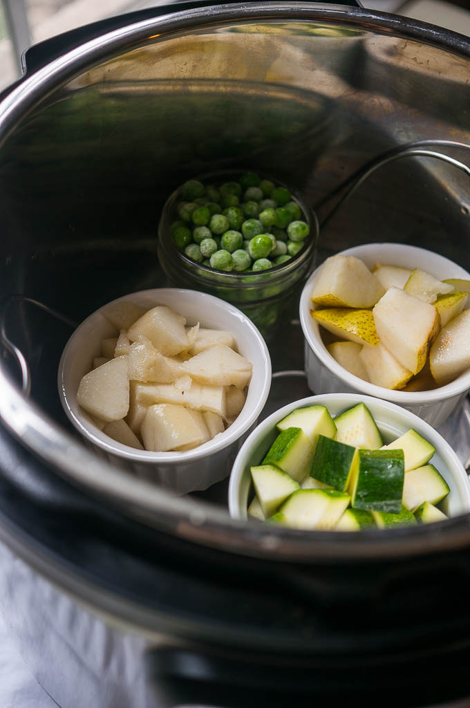 Frozen peas in a glass bowl, pears in a white bowl, zucchini in a white bowl, apples in a white bowl, on a trivet in the instant pot pressure cooker.