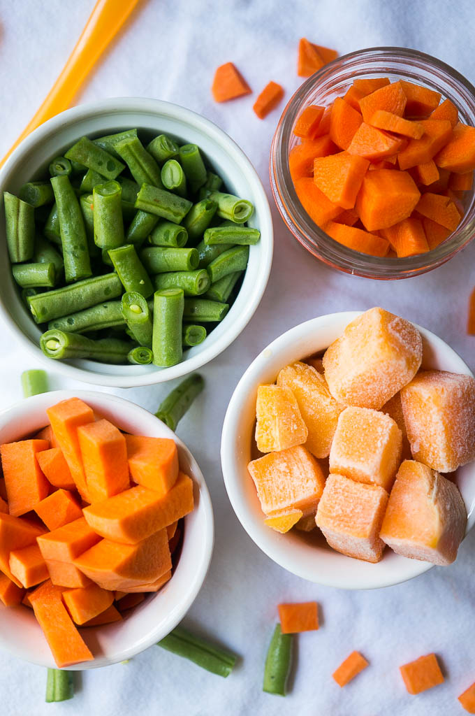 Butternut squash in a white bowl, Sweet Potato in a white bowl, Green beans in a white bowl, Carrots in a glass bowl, Baby spoon, on a white tablecloth.