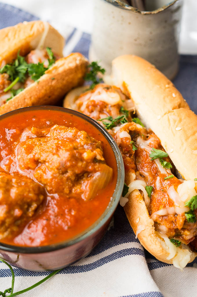 Italian Meatballs topped with parsley and mozzarella cheese in a sesame seed bun on a blue napkin. Italian meatballs and sauce in a glass container. 
