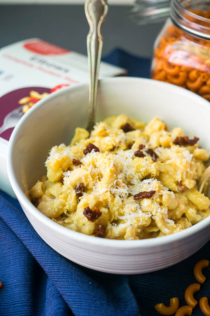 Roasted Cauliflower and sun-dried tomato mac and cheese in a white bowl on a blue napkin.  Box of Modern Table Elbow macaroni complete protein pasta.  Pasta in a glass container.