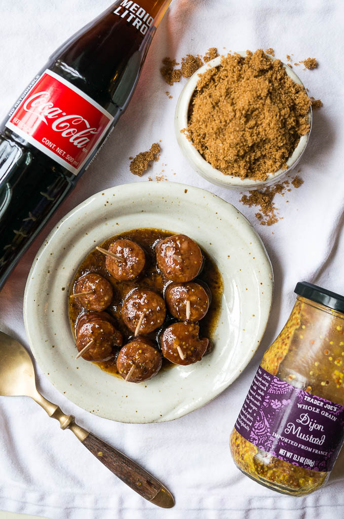 Kielbasa bites on a white speckled plate on a white tablecloth, glass coca-cola bottle, mustard, and brown sugar.