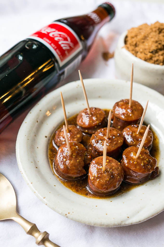 Kielbasa bites on a white speckled plate on a white tablecloth, glass coca-cola bottle, mustard, and brown sugar.