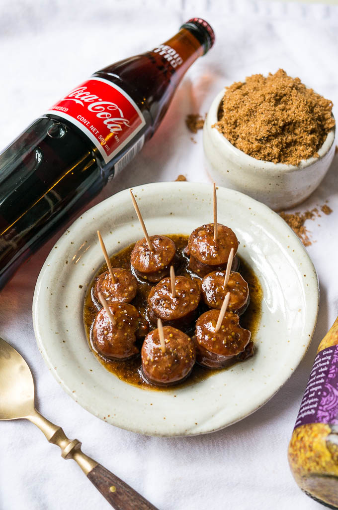 Kielbasa bites on a white speckled plate on a white tablecloth, glass coca-cola bottle, mustard, and brown sugar.