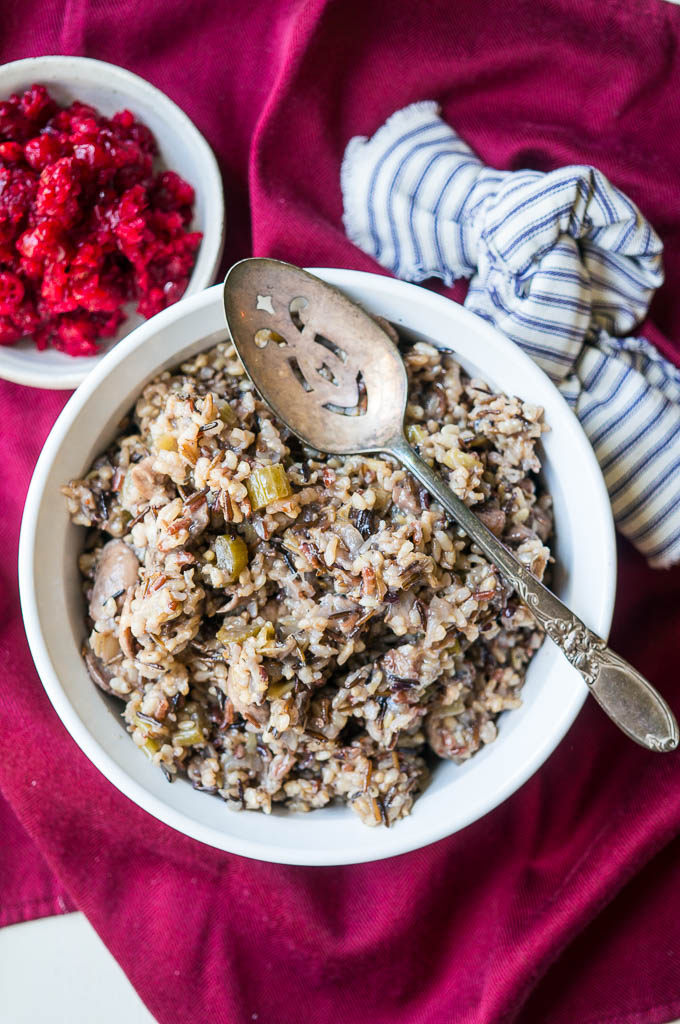 Wild Rice & Mushroom Stuffing in a white bowl with a silver spoon and a blue and white striped napkin and red tablecloth. Cranberry sauce.