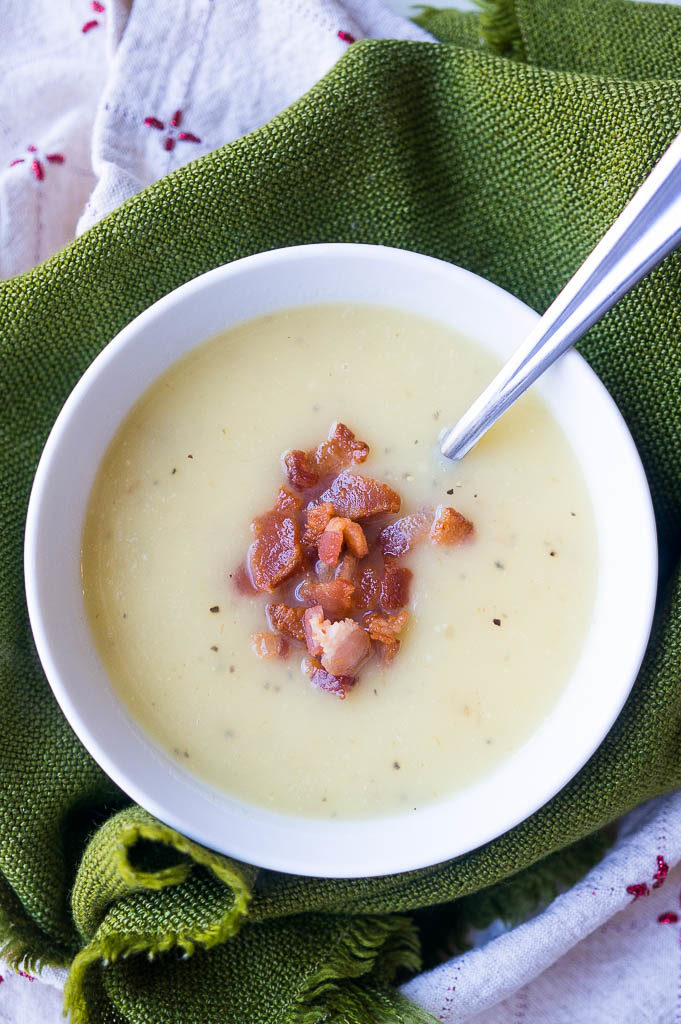 Creamy soup in a white bowl with bacon bits on top and a green background.