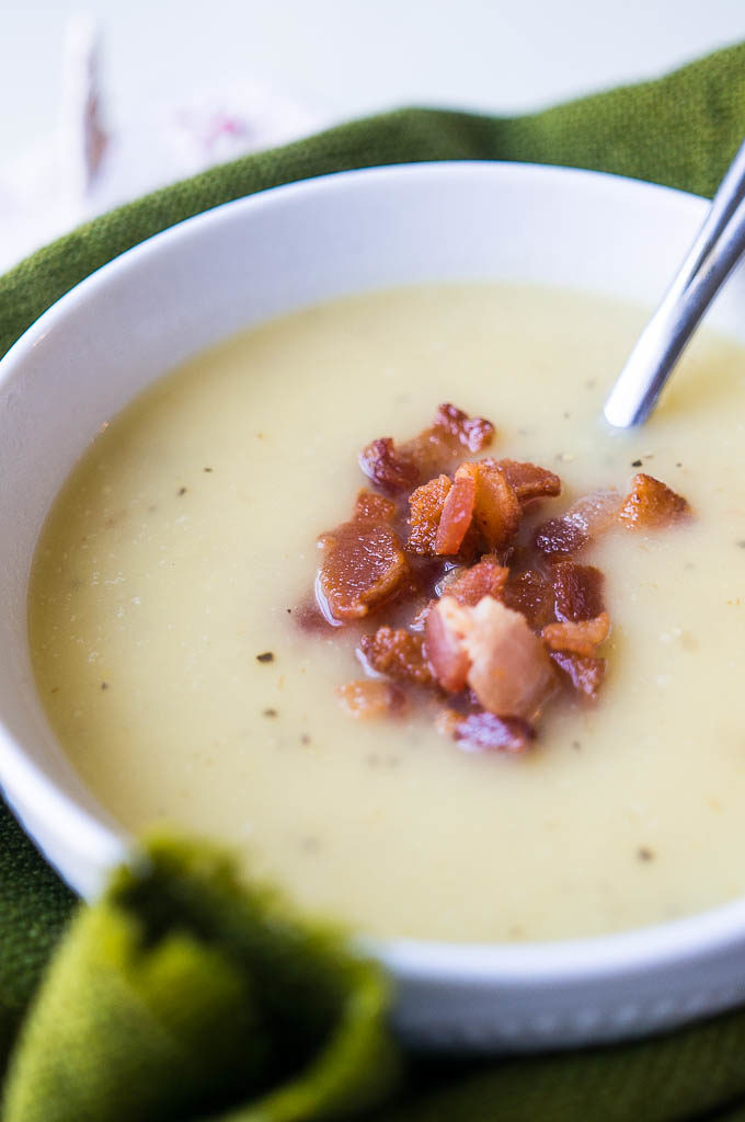 Creamy soup in a white bowl with bacon bits on top and a green background.