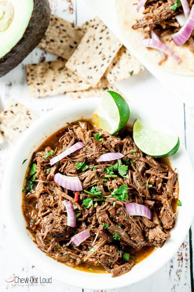 Beef Barbacoa with red onions, cilantro, and lime wedges in a white bowl with tortilla chips and avocado.