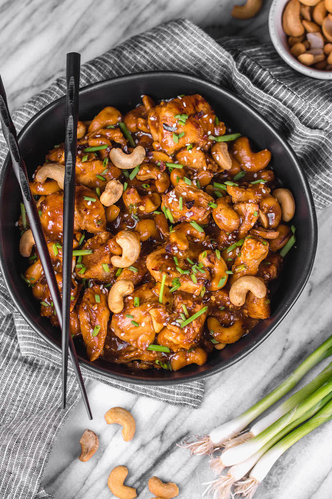 Instant pot cashew chicken garnished with cashews, green onions, and sesame seeds, served in a black bowl with black chop sticks and a grey and white striped napkin on a grey and white marble table.