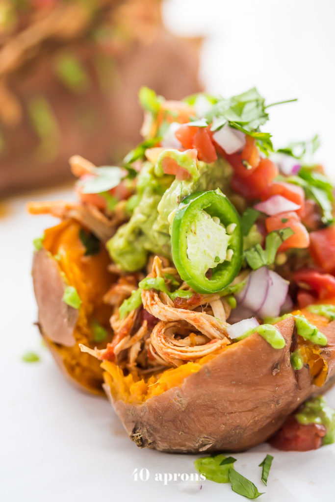 Mexican shredded chicken stuffed sweet potatoes, garnished with guacamole, jalapeños, red onion, cilantro, and tomatoes on a white table.  
